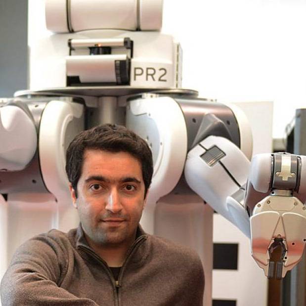photo of Sertac Karaman seated in front of large robot with arms