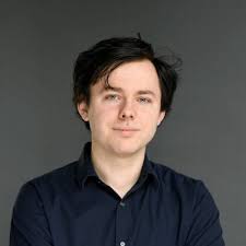 male with dark hair in front of grey background wearing dark blue button up shirt