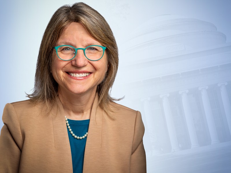 Sally Kornbluth wearing glasses and tan jacket in front of blue graphic of MIT Dome