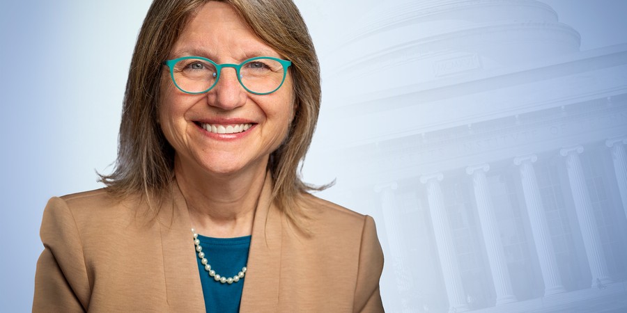 Sally Kornbluth wearing glasses and tan jacket in front of blue graphic of MIT Dome