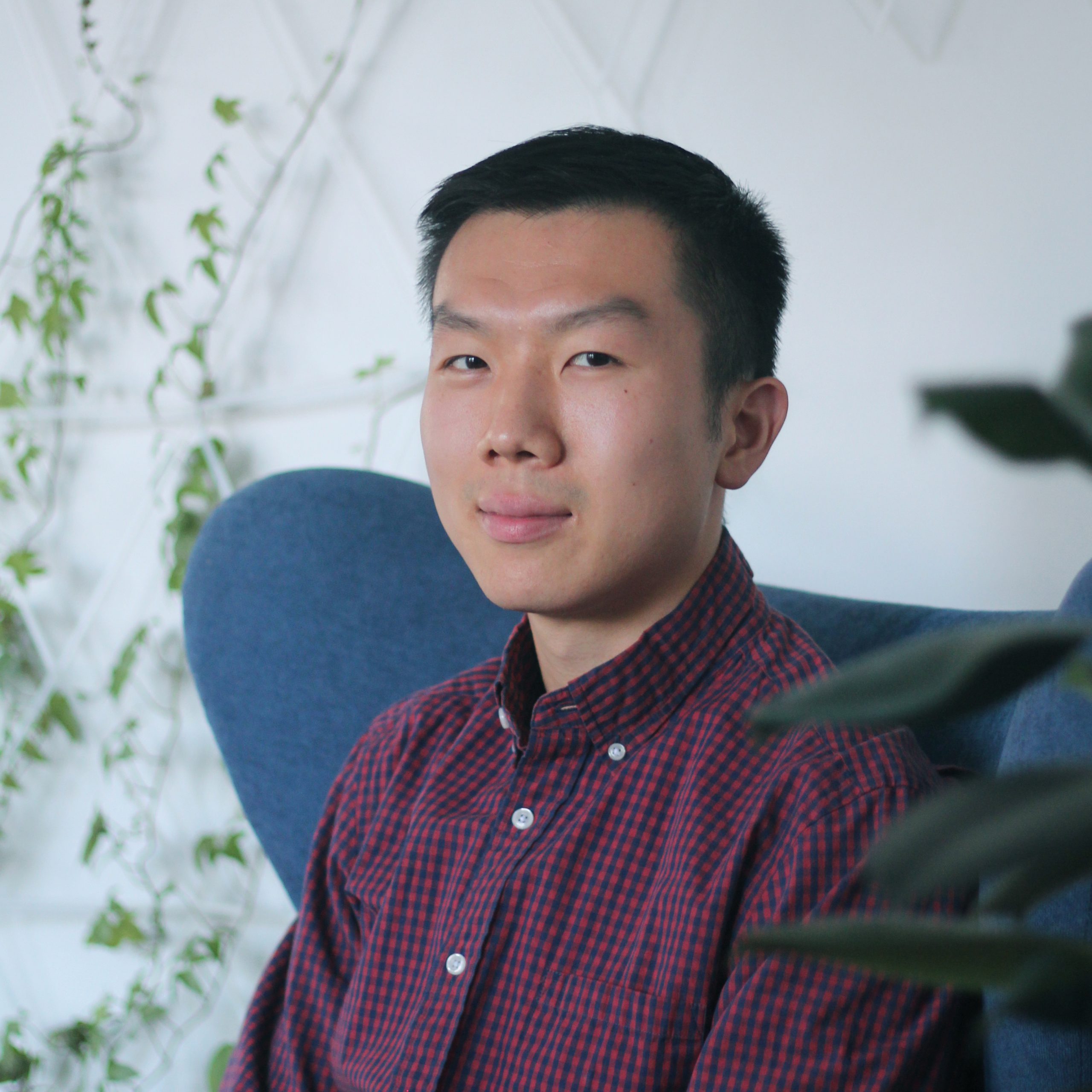 male with dark hair wearing a navy and maroon plaid button up shirt seated in blue chair in front of wall climbing plants