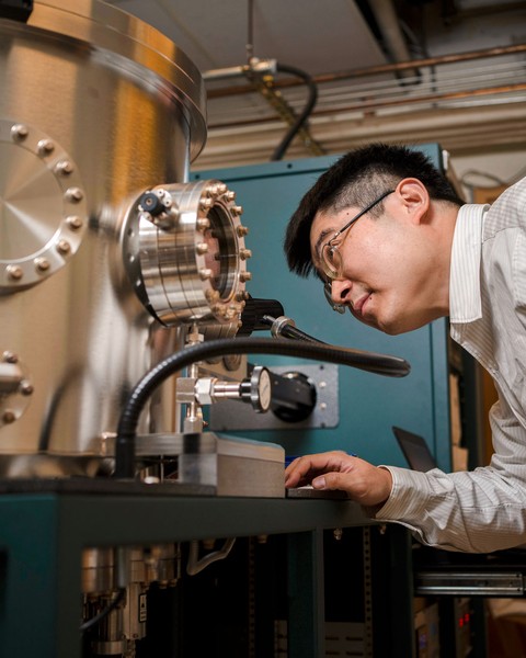 male scientist is bending to look inside window of large silver scientific machinery