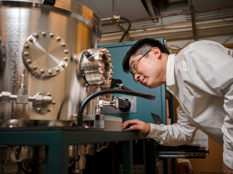 male scientist is bending to look inside window of large silver scientific machinery
