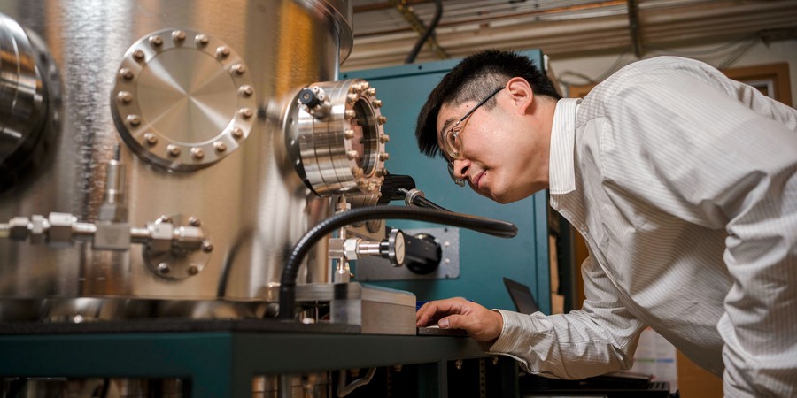 male scientist is bending to look inside window of large silver scientific machinery