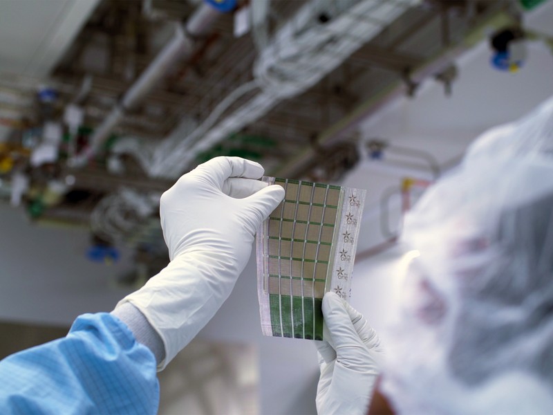 Gloved hands hold a sheet containing 6x5 grid of solar cells.