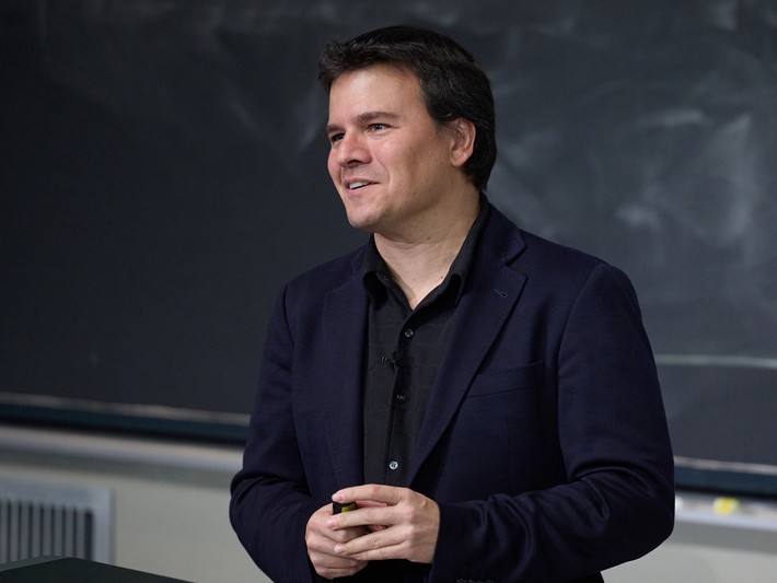 male standing in front of blackboard wearing dark shirt and blue jacket