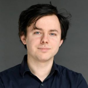 alex sludds a white male with dark brown hair in front of grey background wearing black collared shirt
