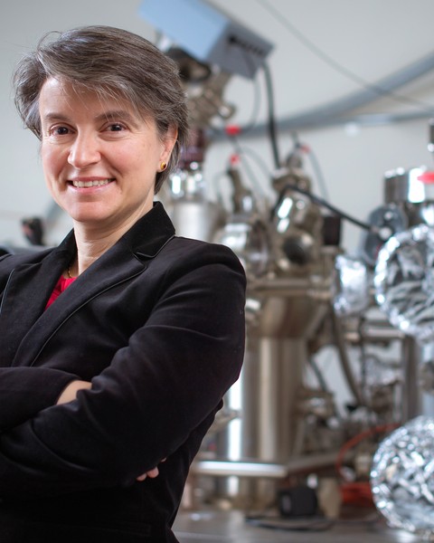 MIT Professor Bilge Yildiz stands with arms folded in her MIT lab with large metallic equipment behind her.