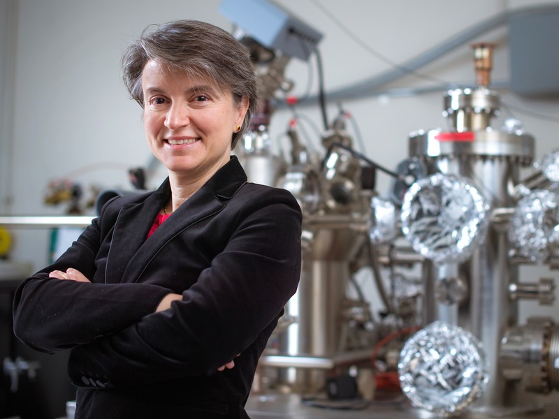 MIT Professor Bilge Yildiz stands with arms folded in her MIT lab with large metallic equipment behind her.