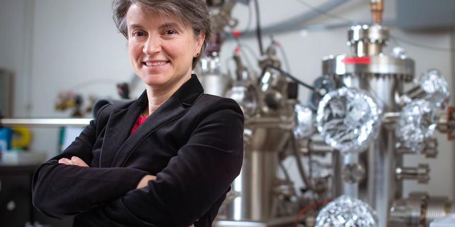 MIT Professor Bilge Yildiz stands with arms folded in her MIT lab with large metallic equipment behind her.