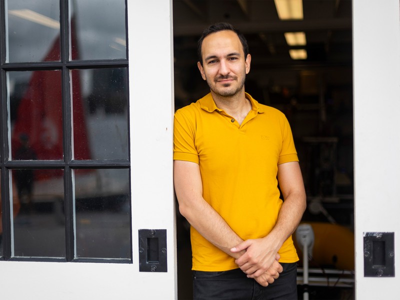Fadel Adib stands between 2 glass panel sliding doors while wearing a yellow shirt and black pants.