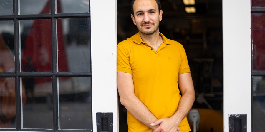Fadel Adib stands between 2 glass panel sliding doors while wearing a yellow shirt and black pants.