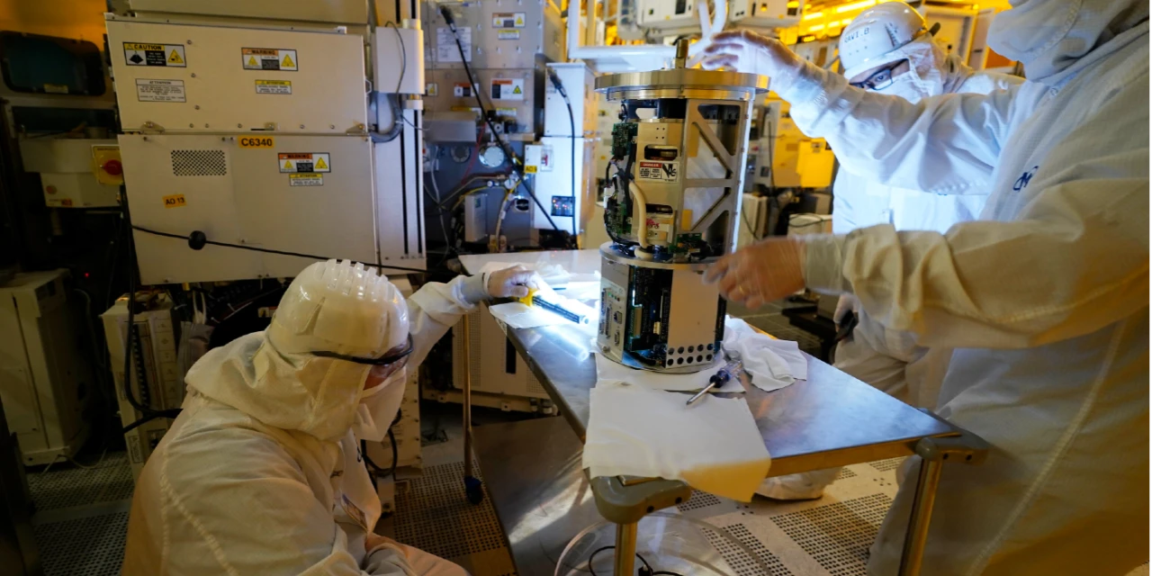 Image Credit: AP Photo/Steve Helber. Technicians wearing white clean room suits inspect a piece of equipment at automotive chip manufacturing plant.