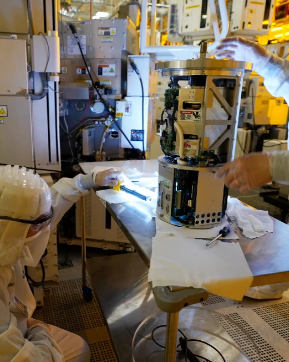 Image Credit: AP Photo/Steve Helber. Technicians wearing white clean room suits inspect a piece of equipment at automotive chip manufacturing plant.