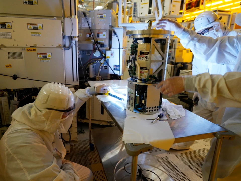 Image Credit: AP Photo/Steve Helber. Technicians wearing white clean room suits inspect a piece of equipment at automotive chip manufacturing plant.