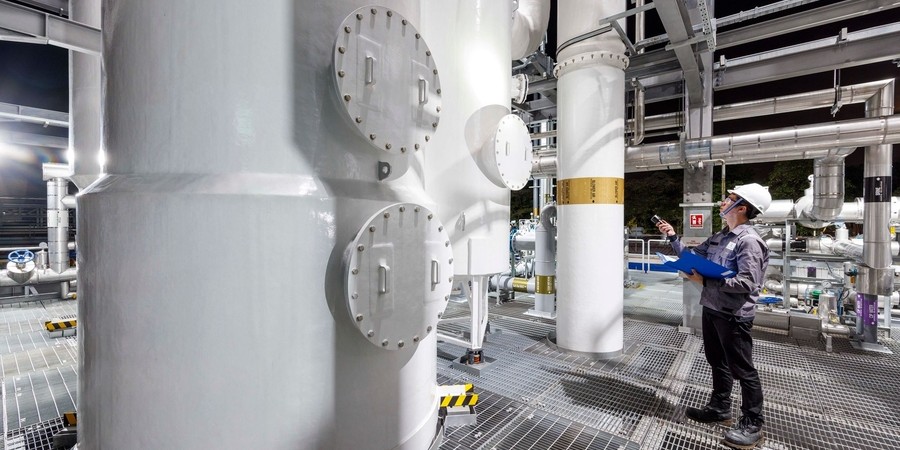 factory scientist wearing hard hat inspecting large white machines using flashlight