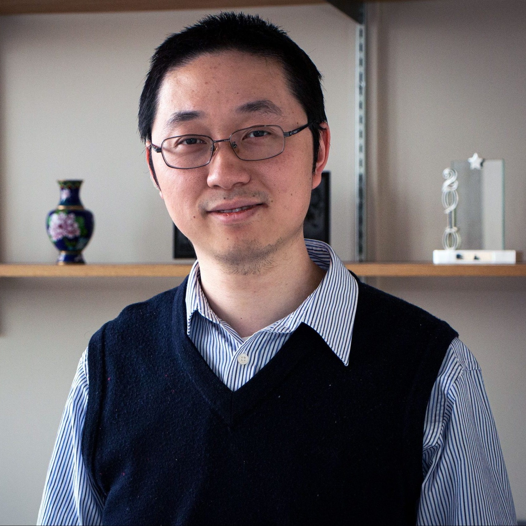 JJ Hu wearing a dark blue vest and a blue and white striped shirt standing in front of wood shelves in his office