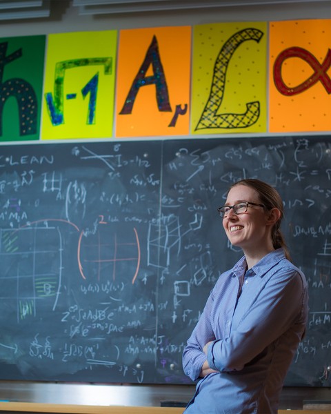 Phiala Shanahan smiles in classroom with blackboard in background, filled with math equations. Colorful posters above of the blackboard show mathematical icons.