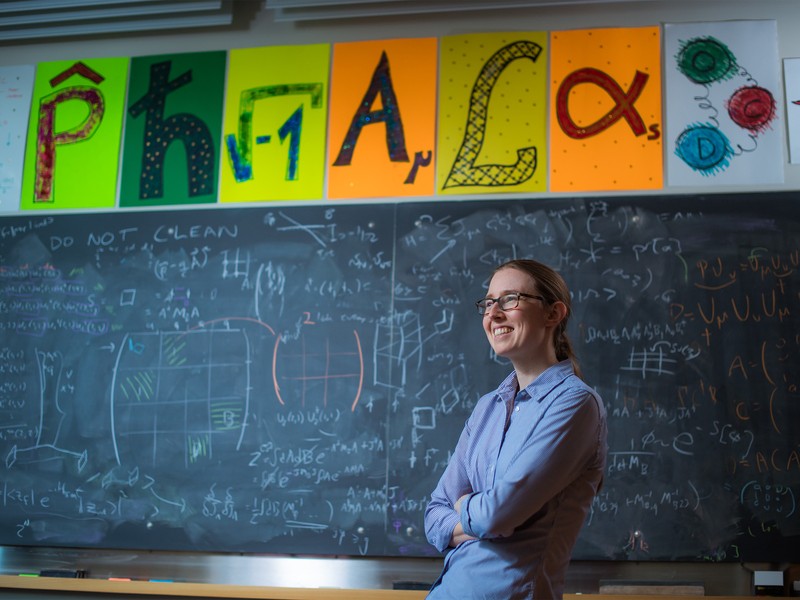 Phiala Shanahan smiles in classroom with blackboard in background, filled with math equations. Colorful posters above of the blackboard show mathematical icons.