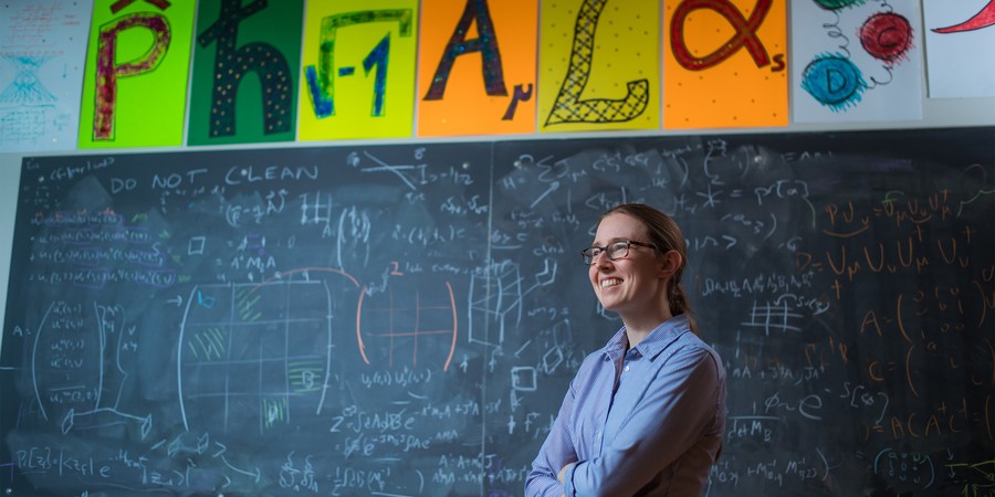 Phiala Shanahan smiles in classroom with blackboard in background, filled with math equations. Colorful posters above of the blackboard show mathematical icons.