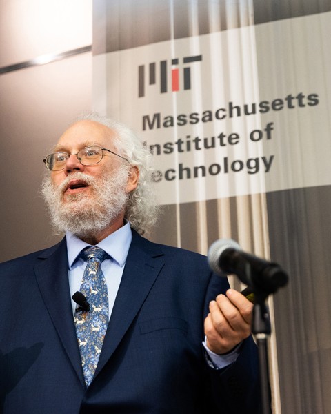 Peter Shor speaking at microphone wearing a blue suit with light blue shirt and patterned blue tie while standing in front of grey background