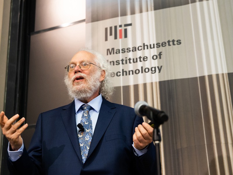 Peter Shor speaking at microphone wearing a blue suit with light blue shirt and patterned blue tie while standing in front of grey background