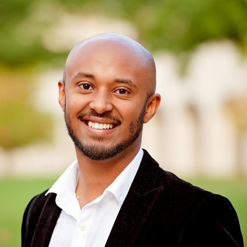male with beard wearing white dress shirt and black jacket standing outside in front of trees