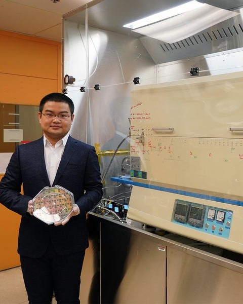Jiadi Zhu wears gloves while holding an octagon-shaped plastic case containing 8-inch wafer. On right, a furnace resembling a deep freezer sits in glass casing.
