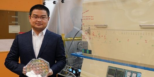 Jiadi Zhu wears gloves while holding an octagon-shaped plastic case containing 8-inch wafer. On right, a furnace resembling a deep freezer sits in glass casing.