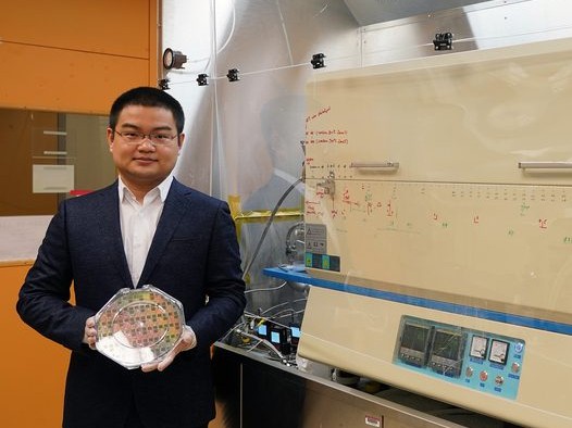 Jiadi Zhu wears gloves while holding an octagon-shaped plastic case containing 8-inch wafer. On right, a furnace resembling a deep freezer sits in glass casing.
