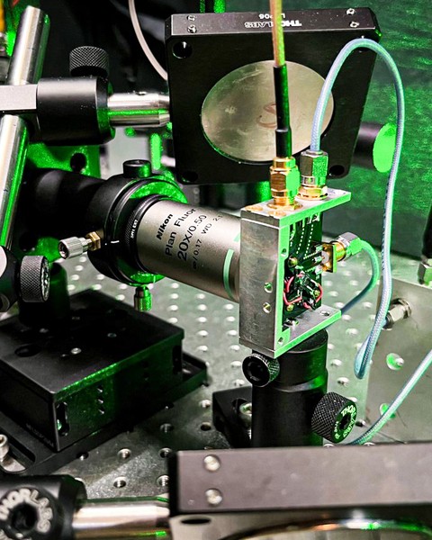 A closeup of the laser equipment shows metallic rods, a circuit board, wires, and lenses, all in a green glow.