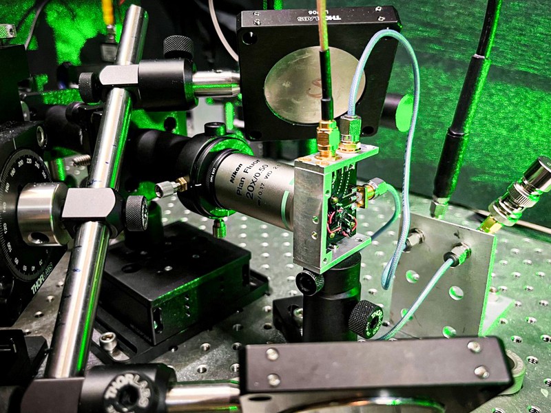 A closeup of the laser equipment shows metallic rods, a circuit board, wires, and lenses, all in a green glow.