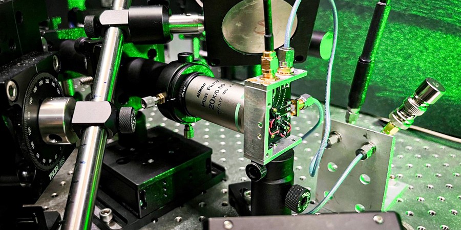 A closeup of the laser equipment shows metallic rods, a circuit board, wires, and lenses, all in a green glow.