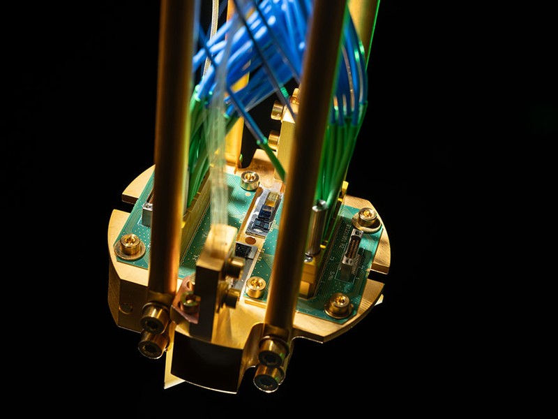 Close-up photo of a quantum repeater module mounted on a gold-plated copper assembly and connected to green printed circuit boards, with optical fibers routed up.