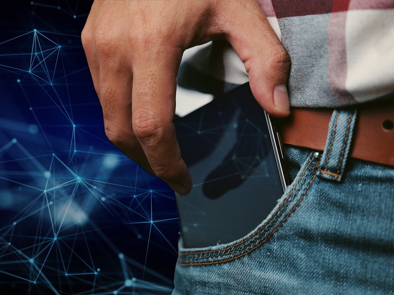 A cell phone peeks out from the pocket of a person wearing jeans, a belt, and a plaid shirt. In the background and on the cell phone’s screen are stylized connected nodes representing a neural network.