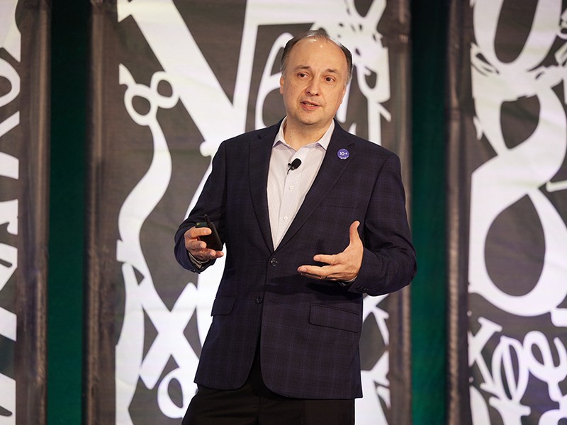 Large white numbers and mathematical symbols appear behind male speaker wearing dark suit and white shirt.