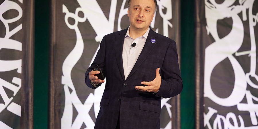 Large white numbers and mathematical symbols appear behind male speaker wearing dark suit and white shirt.