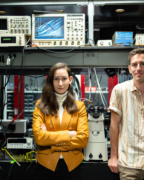 Madeleine Laitz, left, and Dane deQuilettes stand in a lab filled with equipment like computers, microscopes, and oscilloscopes.