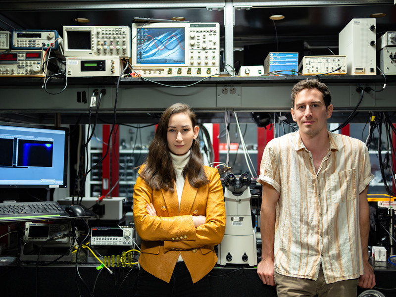 Madeleine Laitz, left, and Dane deQuilettes stand in a lab filled with equipment like computers, microscopes, and oscilloscopes.