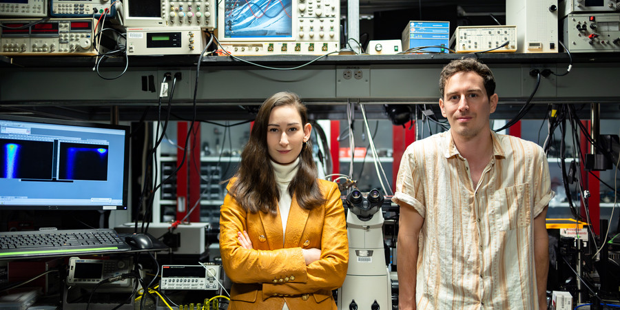 Madeleine Laitz, left, and Dane deQuilettes stand in a lab filled with equipment like computers, microscopes, and oscilloscopes.