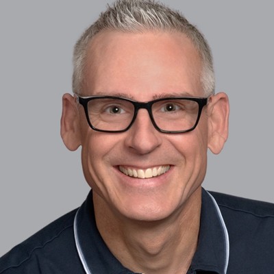 professional headshot photo of a Alan Smith in front of light grey background wearing dark blue polo shirt and black frame glasses