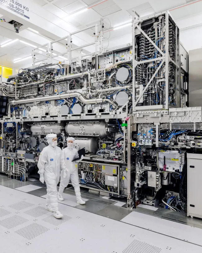 two workers entirely covered in white PPE walk through a factory next to large metal equipment
