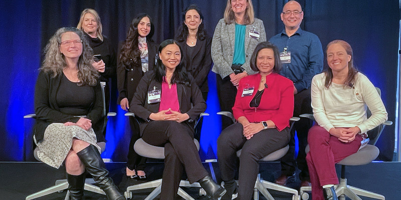 A group photo of eight women and one man in two rows, with back row standing and front seated, on a platform with dark curtains behind them.