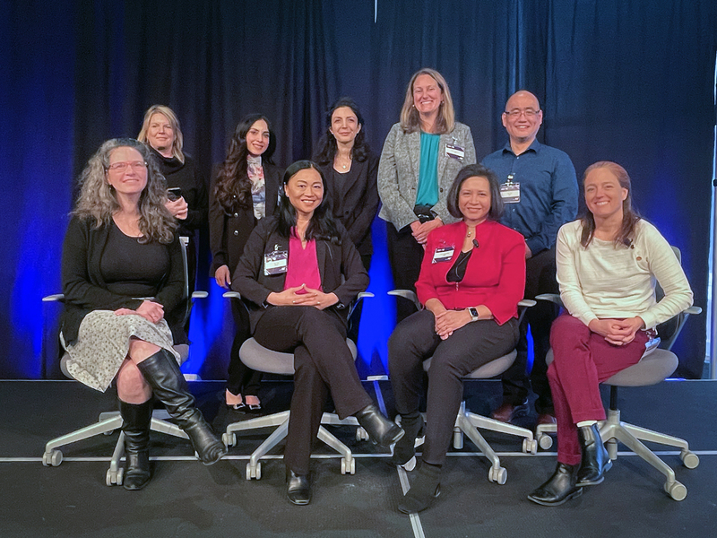 A group photo of eight women and one man in two rows, with back row standing and front seated, on a platform with dark curtains behind them.
