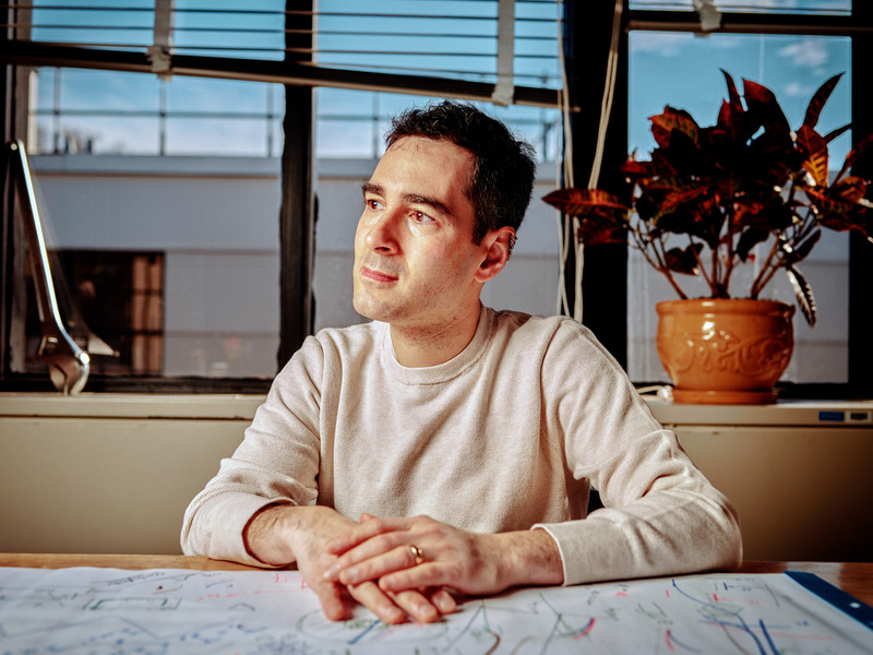 Rafael Jaramillo sits in his office and looks to the side. A large wrench sits on the window sill. The desk is covered in white paper with many drawings and notes on it.