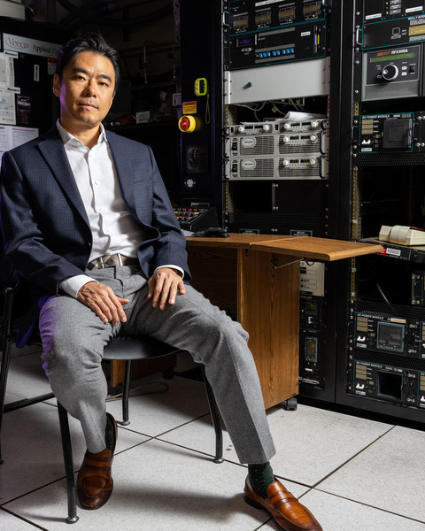 Jeehwan Kim sits on a chair in a dark lab filled with equipment, with a purple light in background.