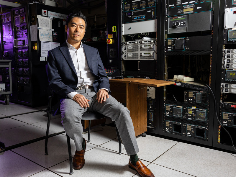 Jeehwan Kim sits on a chair in a dark lab filled with equipment, with a purple light in background.
