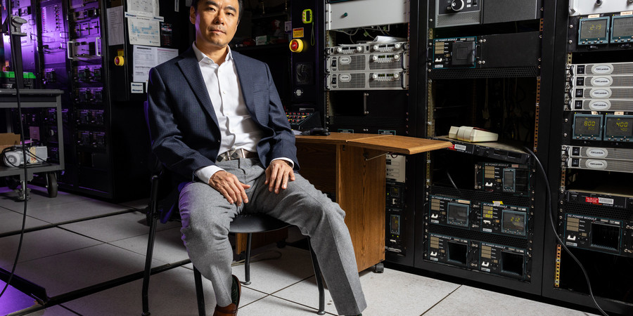 Jeehwan Kim sits on a chair in a dark lab filled with equipment, with a purple light in background.
