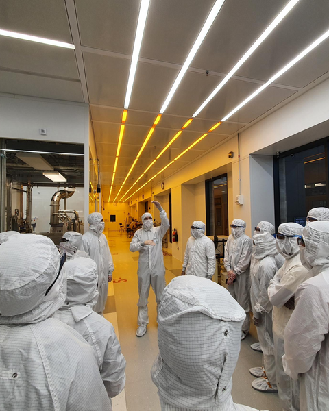 A view from behind of about 15 people dressed head-to-toe in white cleanroom suits, facing another gowned-up individual gesturing as they speak.