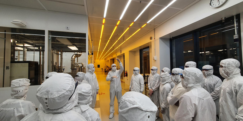 A view from behind of about 15 people dressed head-to-toe in white cleanroom suits, facing another gowned-up individual gesturing as they speak.